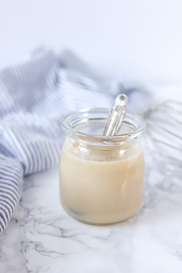 glass jar of homemade sweetened condensed milk with a antique silver spoon coming out. a blue and white stripped towel is in the background