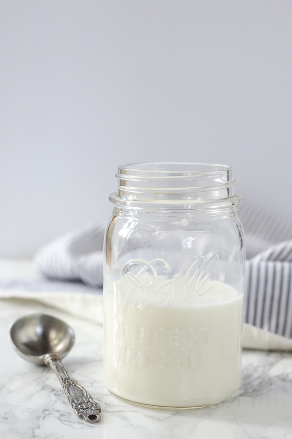 antique mason jar filled with homemade buttermilk with a antique looking measuring spoon to the left and a cream and blue towel in the back