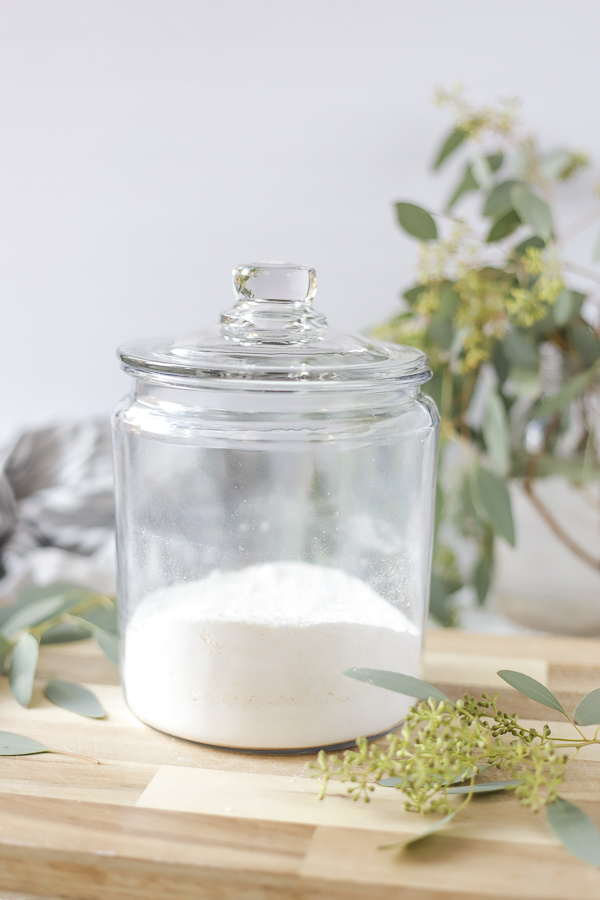 homemade laundry detergent in a glass dish with a lid on a wood cutting board with a mason jar full of eucalyptus