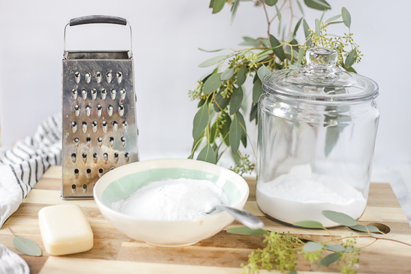 ingredients to make homemade laundry detergent on a wood cutting board with a cheese grater, and eucalyptus leaves behind