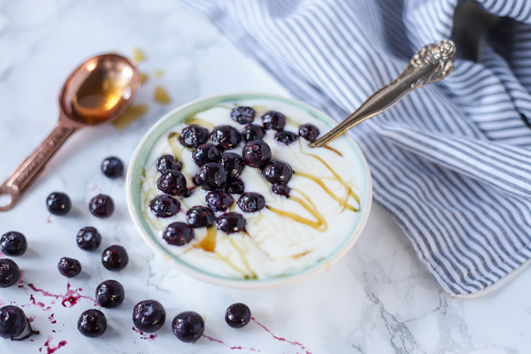 yogurt in a bowl with honey and blueberries on top. Thawed blueberries are sprinkled to the left and a spoonful of honey behind