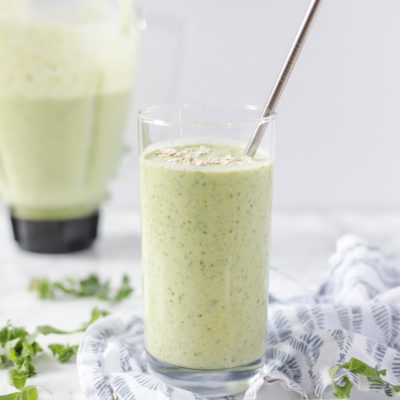 Glass of a tropical green oatmeal smoothie on a blue and white napkin with a blender jar in the background with remaining smoothie in it.