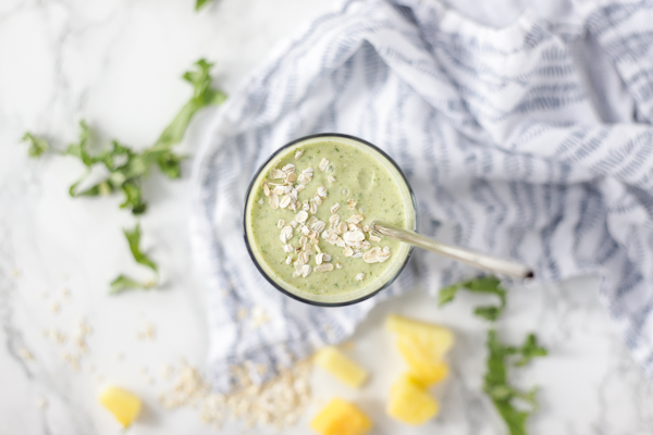 overhead shot of a tropical oatmeal smoothie in a glass with oats on top and a metal straw in the smoothie. The glass is on a white and blue napkin with ingredients spread around