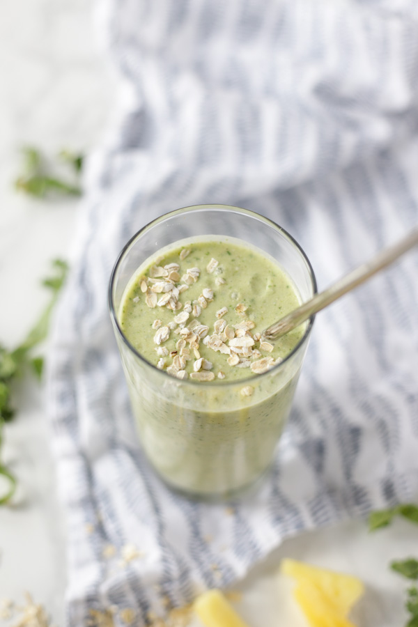 view of the top of a glass filled with a green tropical oatmeal smoothie with a metal straw. The glasses on a blue and white patterned napkin with kale and pineapple spread around