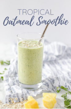tropical oatmeal smoothie in a glass with a metal straw inserted into the smoothie. The glass is on a white and blue patterned napkin with kale, oats, mangos, and pineapples spread around