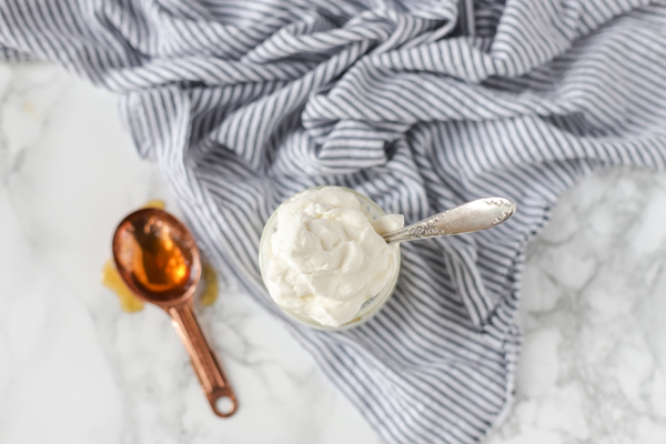 overhead shot of fresh whipped cream in a glass jar on top a stripped towel. A copper spoon with honey sits to the left