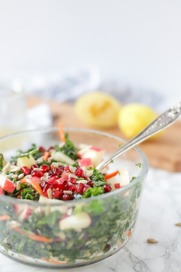 kale salad with carrots, pomegranate, apple slices and sunflower seeds in a glass bowl.