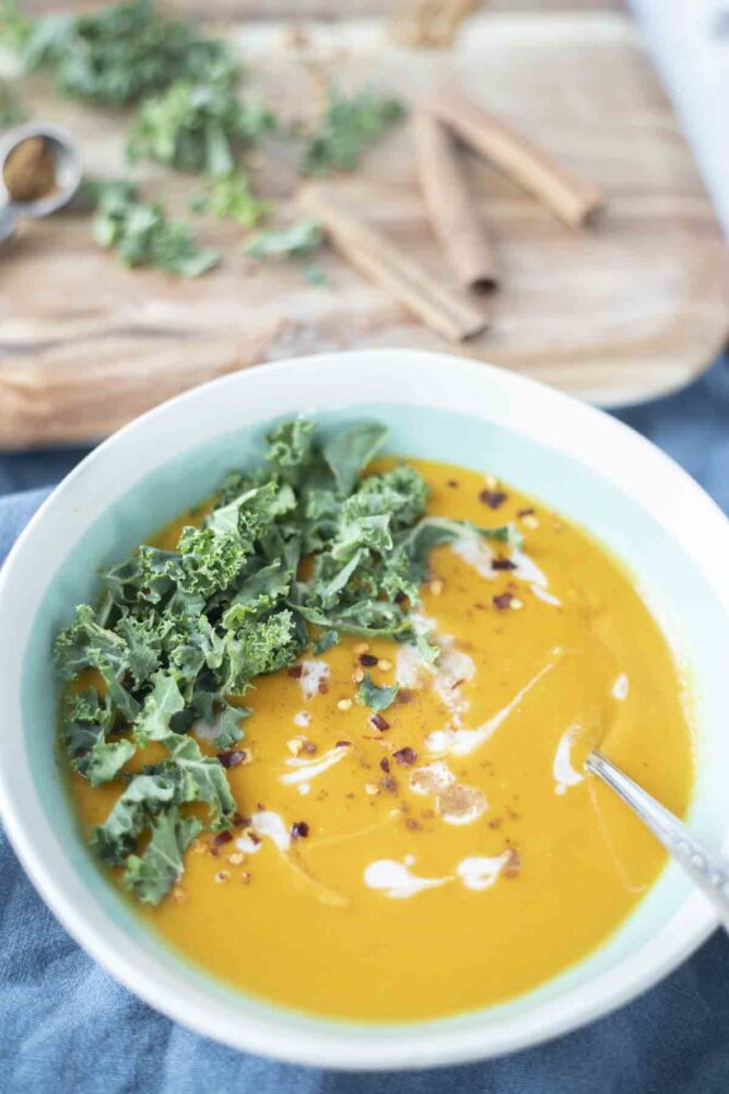 a teal and cream bowl filled with sweet potato and carrot soup topped with chopped kale and coconut cream. There is a cutting board with more kale and cinnamon sticks in the background