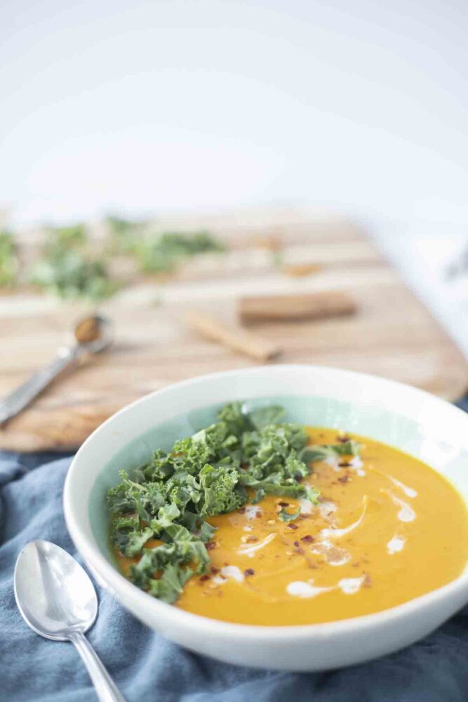 a teal bowl with sweet potato and carrot soup topped with kale and coconut cream on a blue napkin with a spoon on top. A cutting board is in the background
