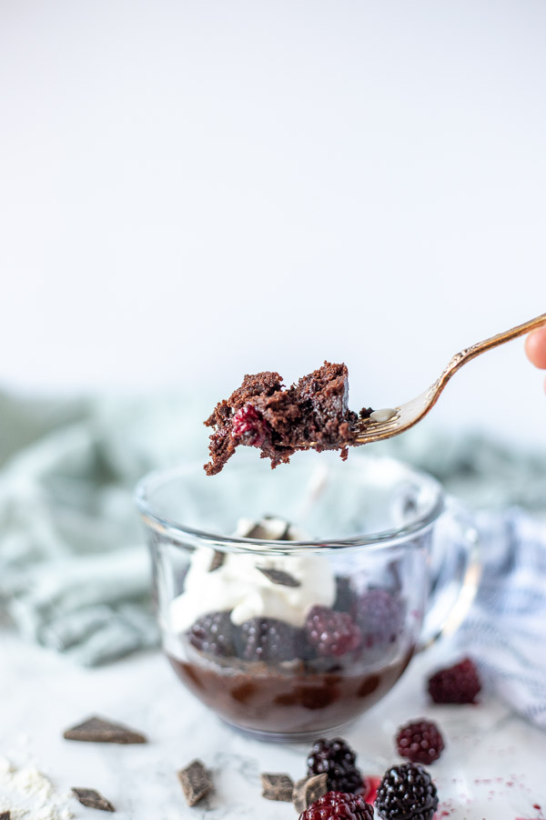 A forkful of blackberry brownie mug cake above the mug full of cake topped with whipped cream and chocolate chips