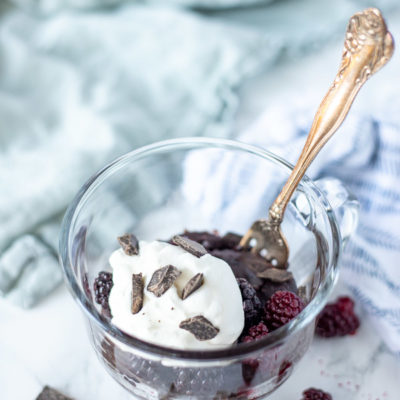 blackberry brownie mug cake in a glass mug. Brownie topped with berries, whipped cream, and chocolate chips. And antique fork is in the mug