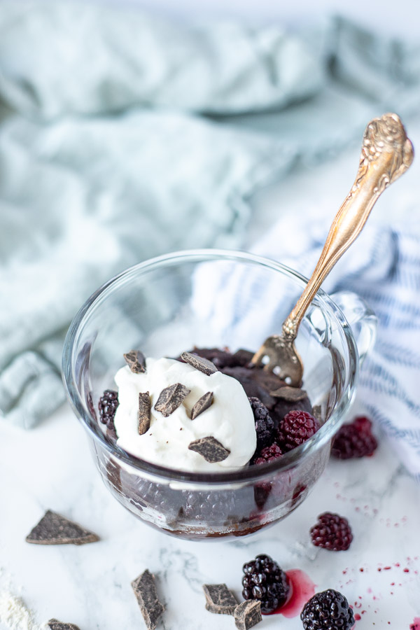 blackberry brownie mug cake in a glass mug. Brownie topped with berries, whipped cream, and chocolate chips. And antique fork is in the mug