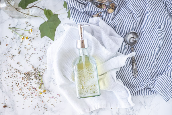 homemade body wash laying o a white towel on a white countertop with dried florals surrounding the bottle