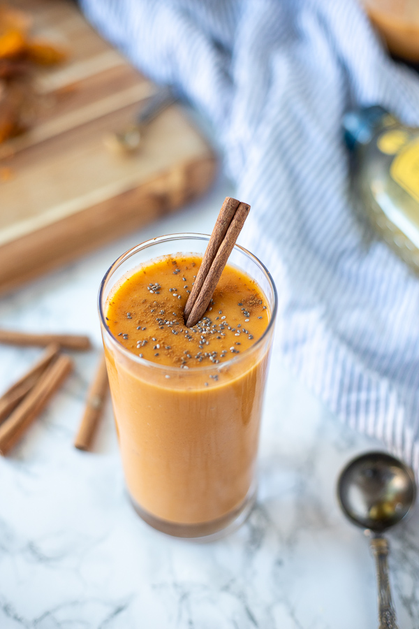 Sweet potato smoothie with chia seeds sprinkled on top and a cinnamon stick in a tall glass. Ingredients and a striped towel in the background
