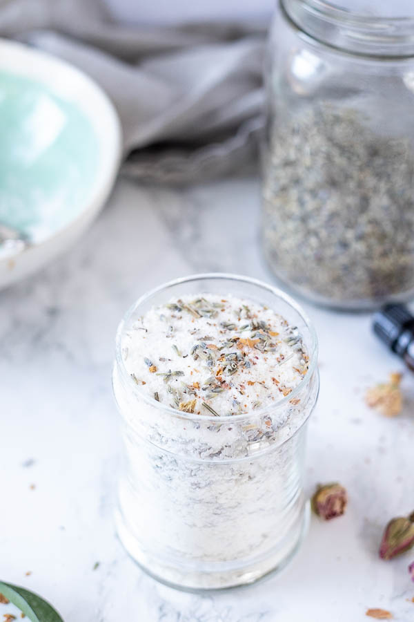 DIY bath salts with dried flowers in a glass jar. A bowl and a jar of dried lavender in the background