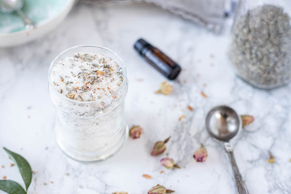 jar of DIY bath salts with dried flowers. Essential oil bottle, measuring spoon, and more dried flowers are spread around the jar