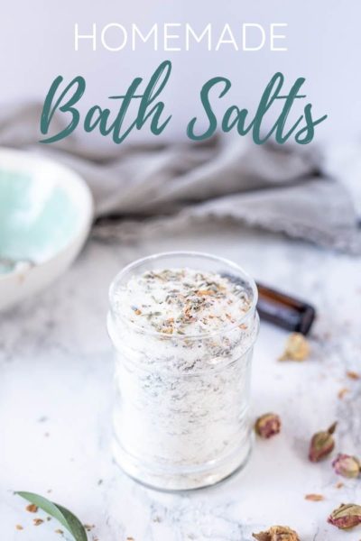 homemade bath salts in a glass jar with herbs on top. Dried herbs and flowers are around the jar