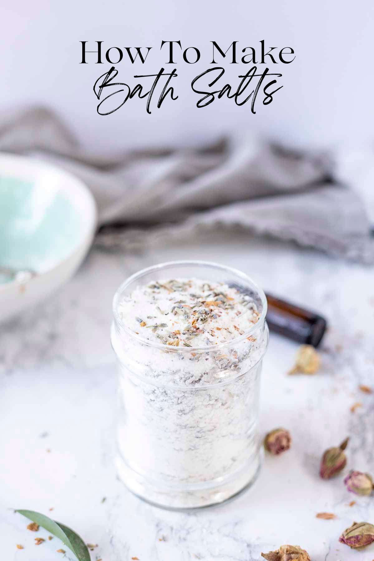homemade bath salts in a glass jar with herbs on top. Dried herbs and flowers are around the jar.