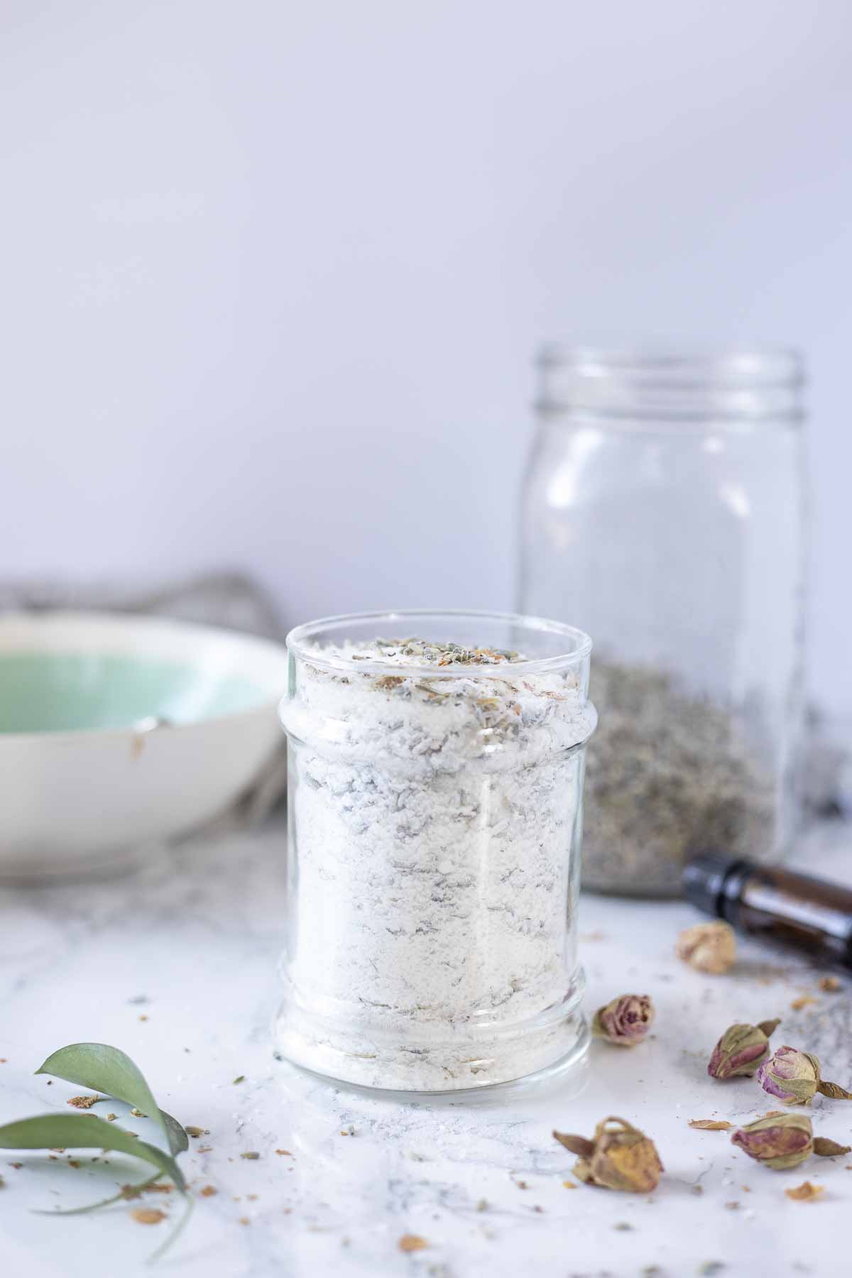 Sideview of a glass container of bath salts with dried flowers on a marble countertop with more dried flowers and glass containers.