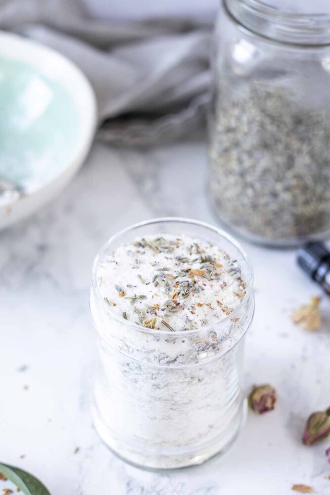 DIY bath salts with dried flowers in a glass jar. A bowl and a jar of dried lavender in the background