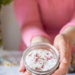 women wearing a pink shirt holding a small jar of homemade foot soak topped with dried roses