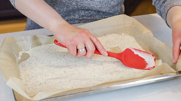 person with a red spatula smoothing out sourdough cracker batter on a parchment lined cookie sheet