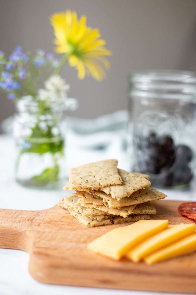 a wood cutting board with homemade sourdough crackers stacked up and cheese slices. Olives in a jar in the background