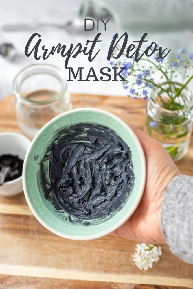 a hand holding a armpit detox mask in a teal and cream bowl. A wood cutting board with ingredients in the background