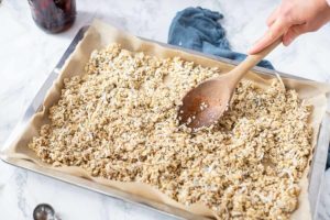 coconut granola evenly spread out on a baking sheet with a wooden spoon, ready for baking