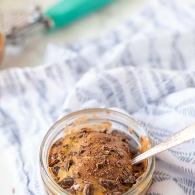short glass dish of peanut butter and ice cream topped topped with peanut butter and chocolate shavings. An antique spoon is in the ice cream with an teal ice cream scoop in the background