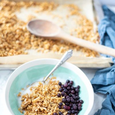 coconut granola with yogurt and blueberries in a teal and cream colored bowl. A pan of freshly baked coconut granola in the background