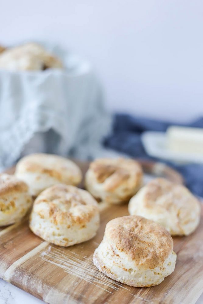 6 sourdough biscuits on a wood countertop with a  lined bowl of biscuits in the background