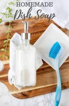 homemade dish soap in a glass soap pump laying on a white wash cloth on a wood cutting board with a white dish to the right with a blue dish scrubber