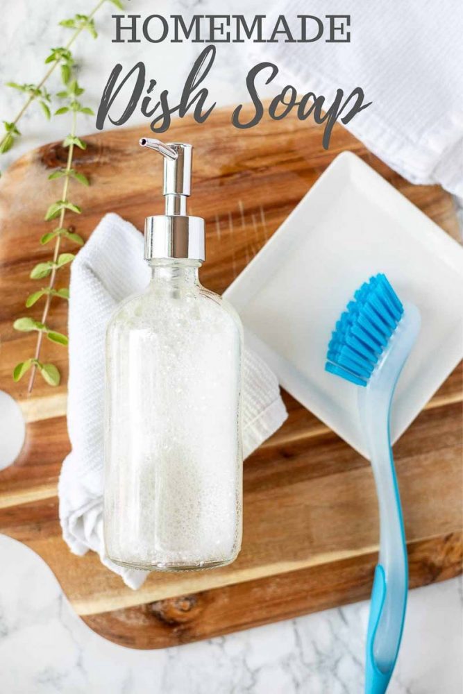 homemade dish soap in a glass soap pump laying on a white wash cloth on a wood cutting board with a white dish to the right with a blue dish scrubber