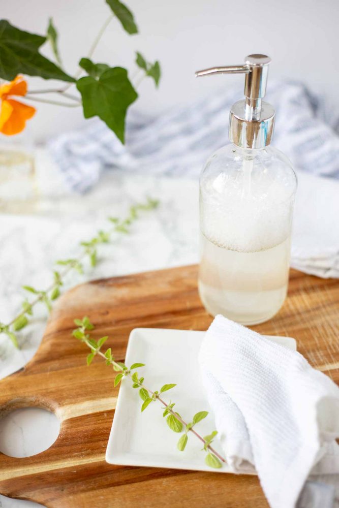 wood cutting board with a white plate and a rag on top and natural dish soap in a glass bottle in the back