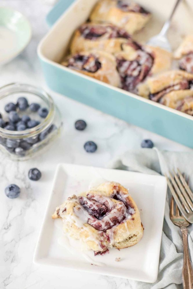 sourdough blueberry sweet roll on a white plate with blueberries and baking dish or rolls in the background