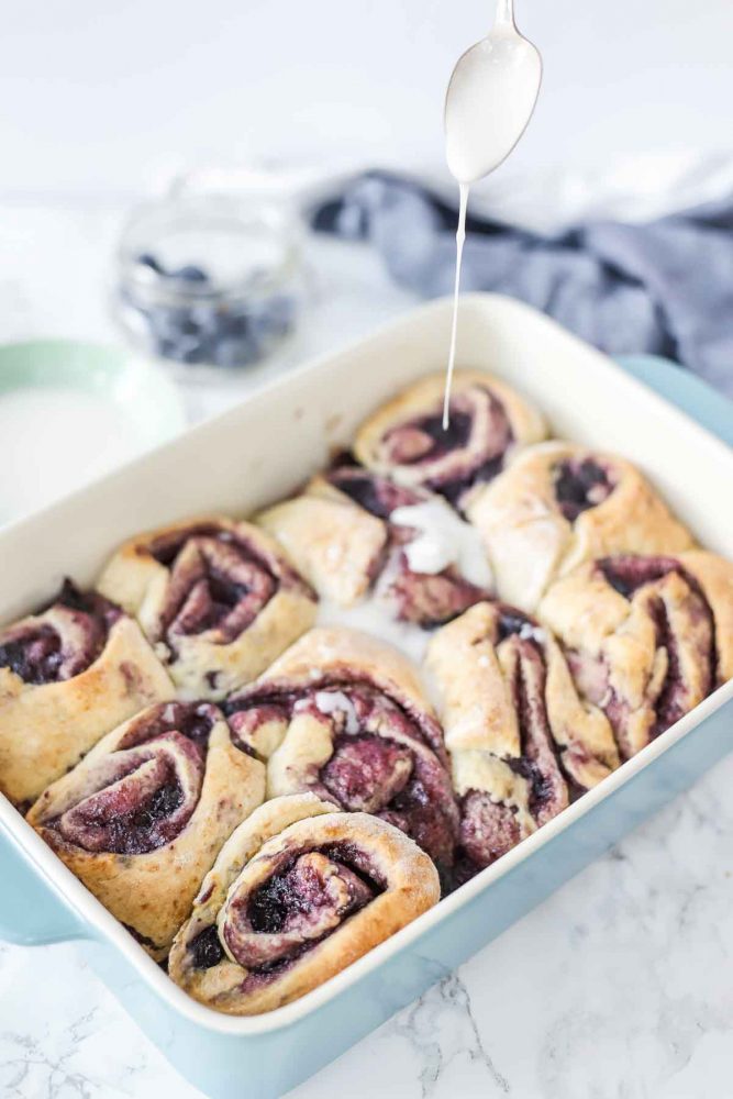 icing being drizzled over cooled blueberry rolls in a baking dish