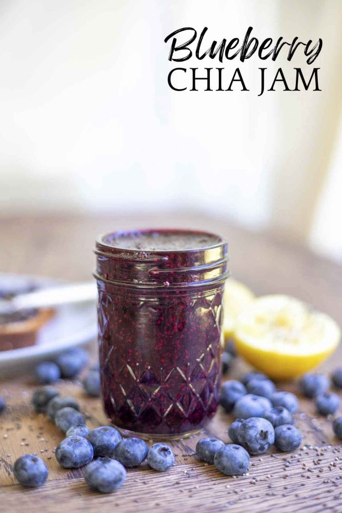 jar of blueberry chia jam on a wood table surrounded by fresh blueberries, chia seeds, and lemon slices.