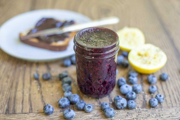 jar of blueberry freezer jam on a wood table with fresh blueberries and sliced lemons surrounding the jar. A plate of toast smothered in jam is in the background