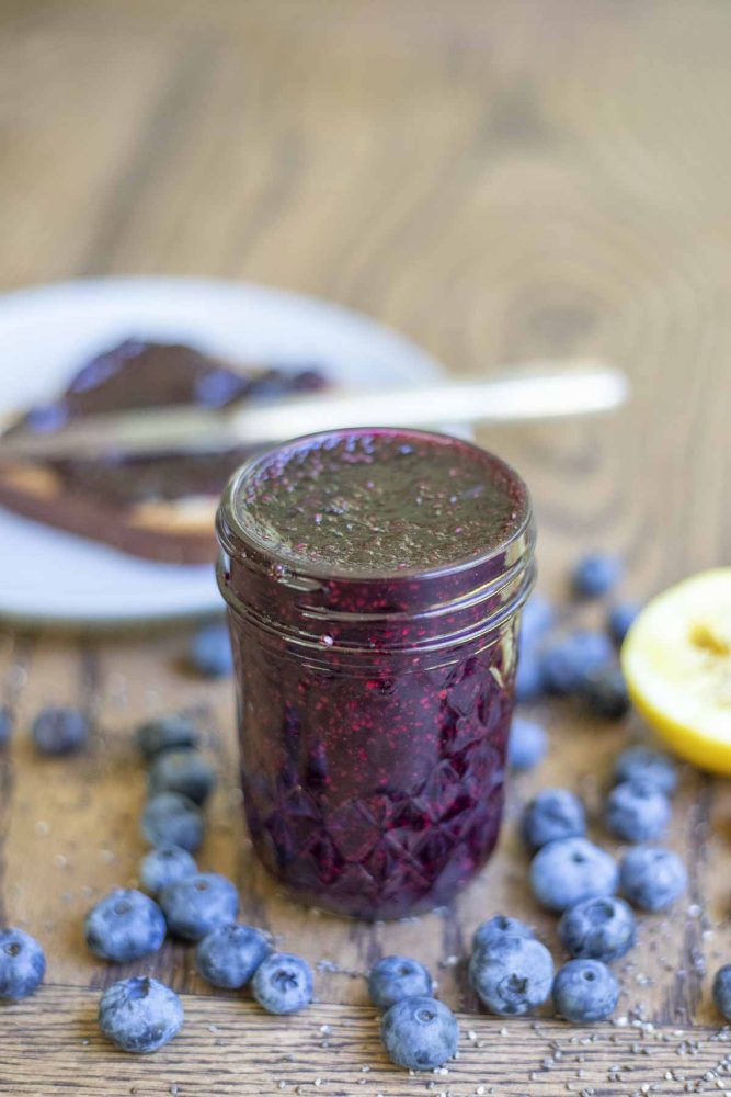 jar of blueberry chia jam in a small mason jar surrounded by fresh blueberries. A plate with toast with jam and a knife in the background