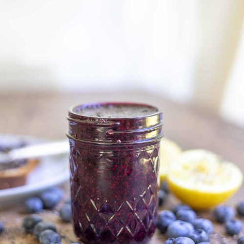 jar of homemade blueberry chia jam on a wood table with blueberries spread around and sliced lemons