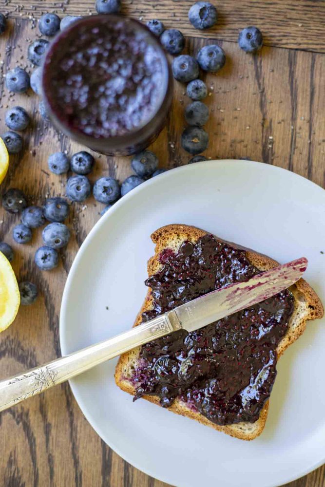 toast smothered with blueberry chia jam with a knife laying on the toast on a white plate. Blueberries and more jam surround the plate