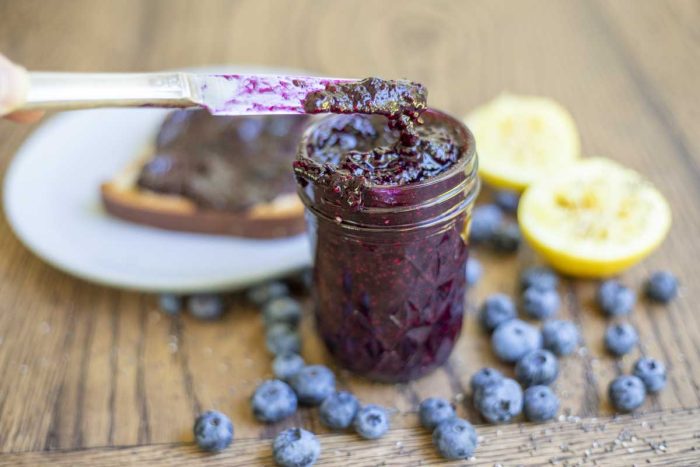 knife scooping out blueberry jam out of a small mason jar. The jar is surrounded by blueberries, lemons, chia seeds, and a plate with toast