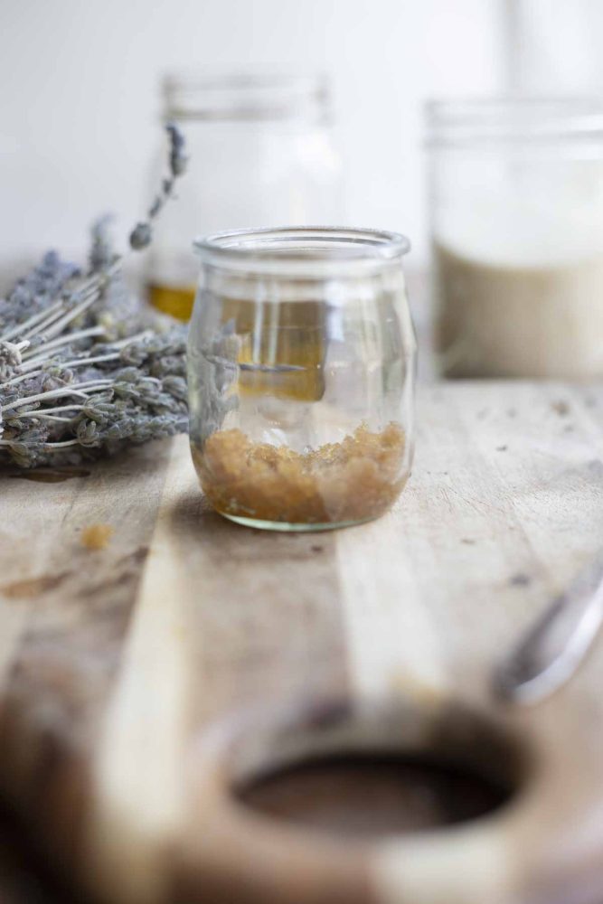 jar of brown sugar lip scrub on a wooden cutting board with a bundle of lavender and jars of oil and sugar in the background