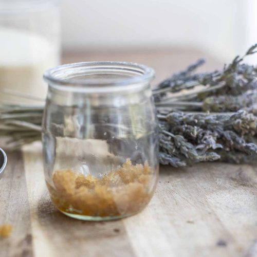 jar of lip sugar scrub on a wood countertop with a bundle of lavender and a glass of sugar in the background