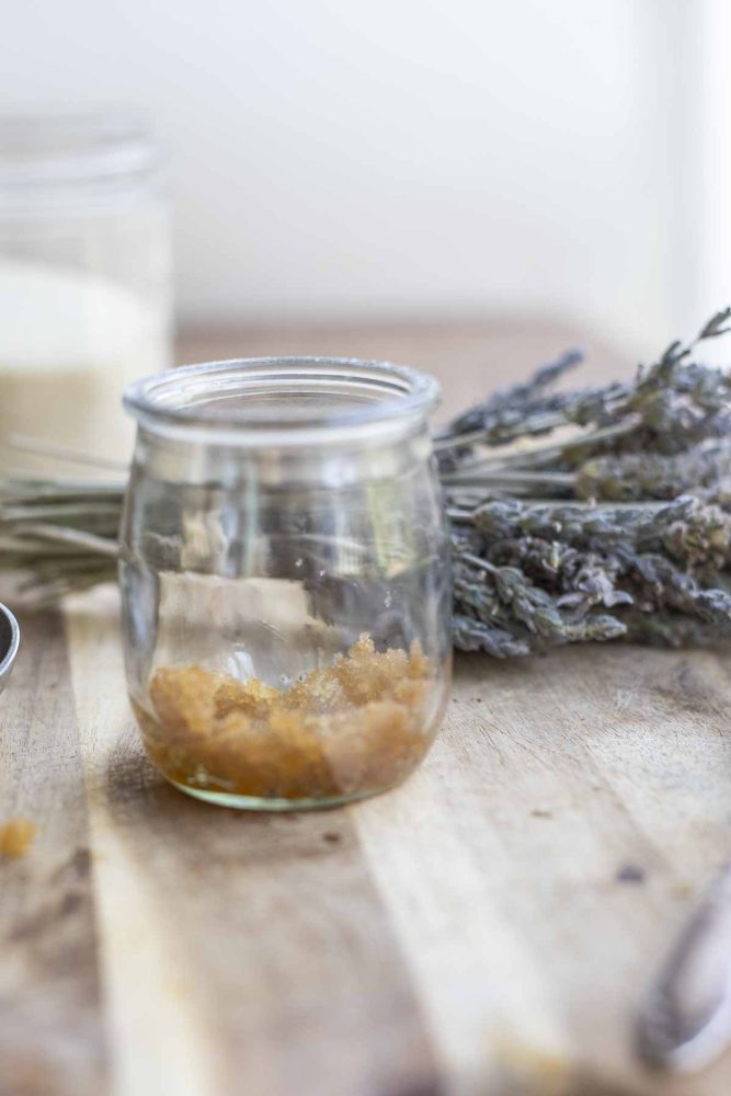 jar of lip sugar scrub on a wood countertop with a bundle of lavender and a glass of sugar in the background