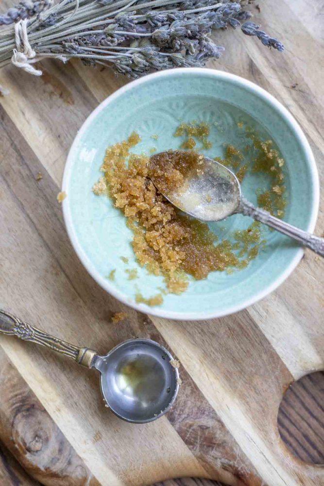 teal bowl of brown sugar lip scrub with a spoon on a wood cutting board. A bundle of lavender and a tablespoon measuring spoon surrounds the bowl