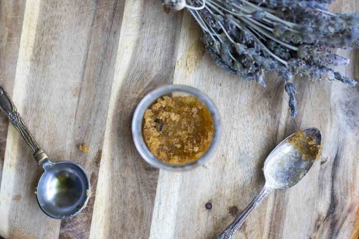 overhead photo of lip scrub in a glass jar with a measuring spoon and a tea spoon around the jar. A bundle of lavender is in the background