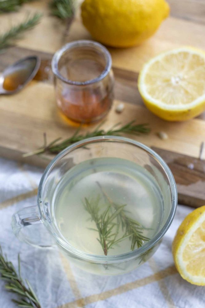 rosemary tea with fresh sprigs of rosemary in a glass mug with a cutting board with a jar of honey, lemon slices, and rosemary in the background