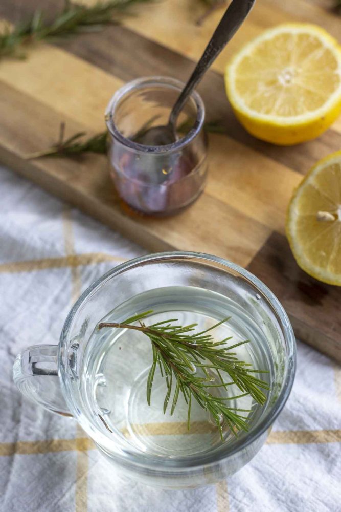 fresh sprigs of rosemary in a glass mug on a white and yellow towel with a wood cutting board with sliced lemons and a jar of honey in the background